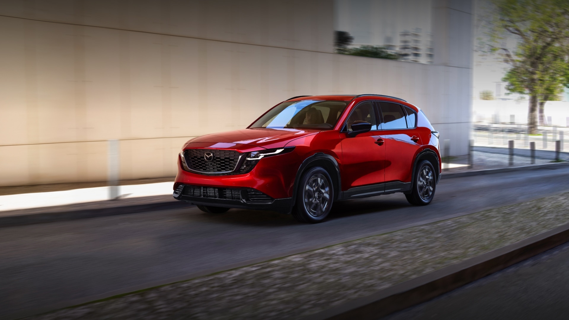A red Mazda CX-5 SUV driving along a city street with a modern backdrop.