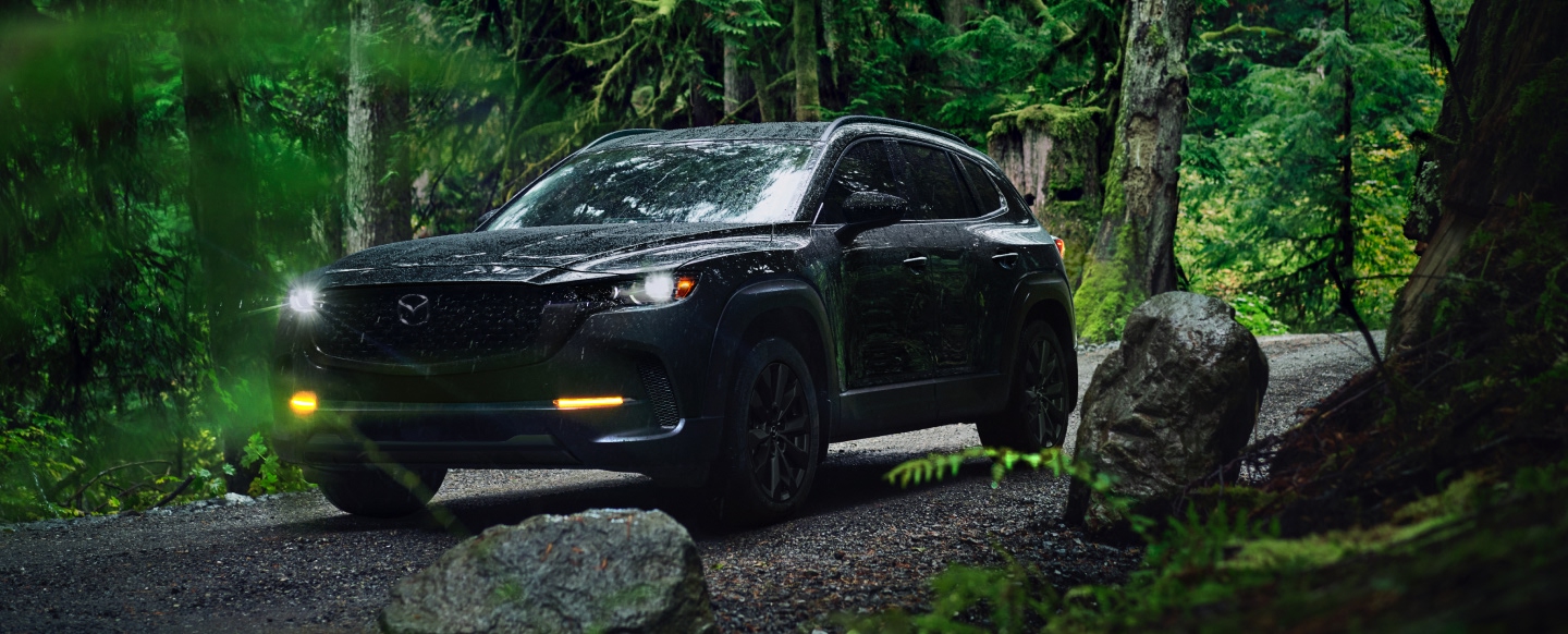 Black Mazda CX-50 SUV driving through a rainy forest trail surrounded by trees and rocks.