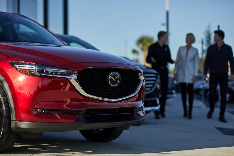 A shiny red Mazda car is in the foreground, with three people walking in the background.