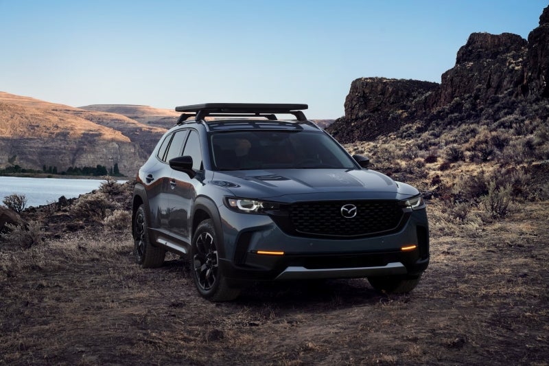 A dark gray Mazda CX-50 parked on rugged terrain with a body of water and rocky hills in the background.