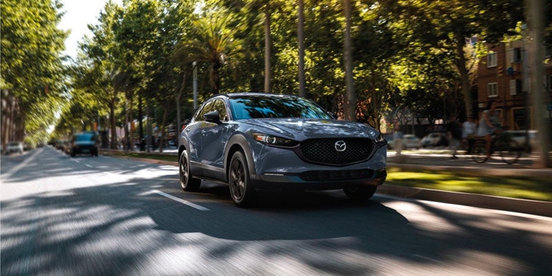 A gray Mazda CX-30 drives down a tree-lined street with motion blur.