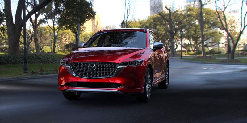 A red Mazda CX-5, viewed from the front, is parked on a paved road with trees and buildings in the background.