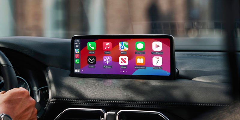 A person's bare arm and hand holding the steering wheel of a car, with the Apple CarPlay screen featuring several app icons in the background.
