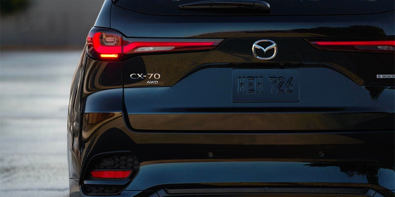 Rear view of a dark blue Mazda CX-70 SUV, showing a red taillight and the Mazda logo.
