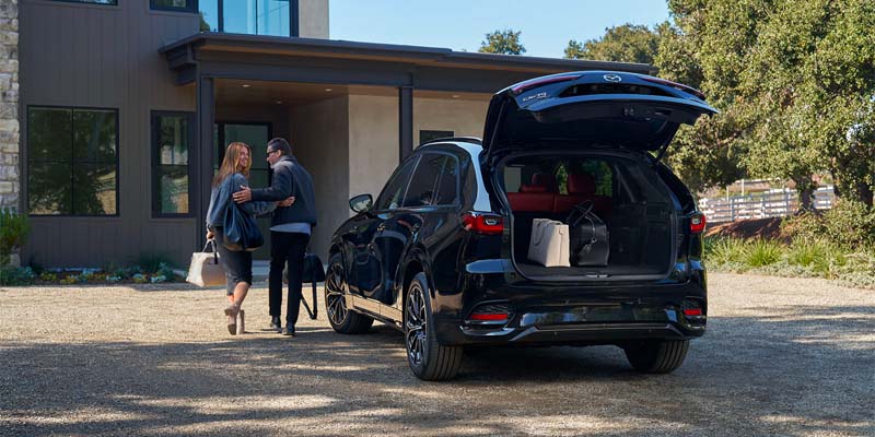 A couple walks toward a black SUV with its trunk open, revealing two bags. They're on a gravel driveway in front of a modern house.