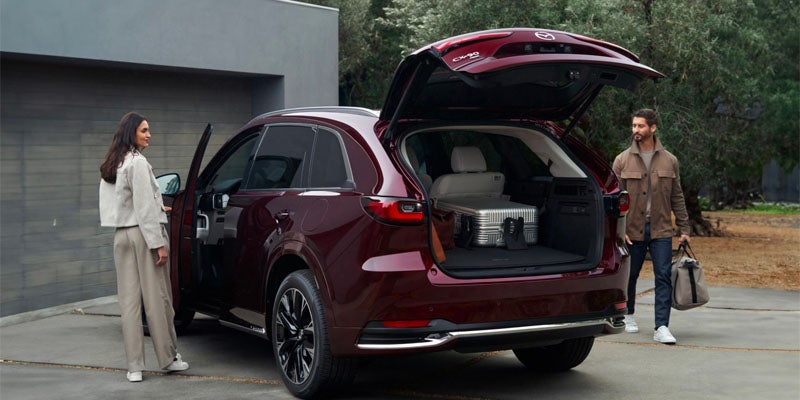A couple loading luggage into the open trunk of a burgundy Mazda CX-90. The woman stands by the driver's side.