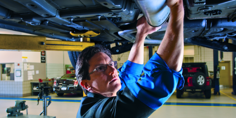 A man in glasses and a blue shirt works on the underside of a car on a lift in a garage.
