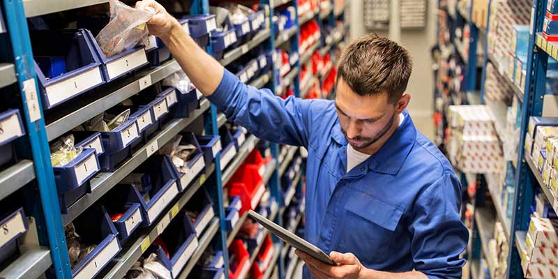 A man in a blue uniform takes an item from a shelf in a warehouse while looking at a tablet.