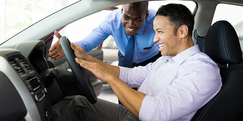 A car salesman smiles as a man sits in a new car, hands on the steering wheel, happily looking at the dashboard.